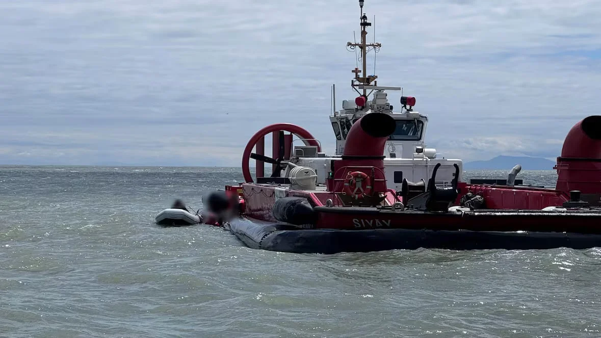 A large red and white hovercraft vessel sits in choppy open water with a smaller inflatable boat secured alongside it under a cloudy sky.
